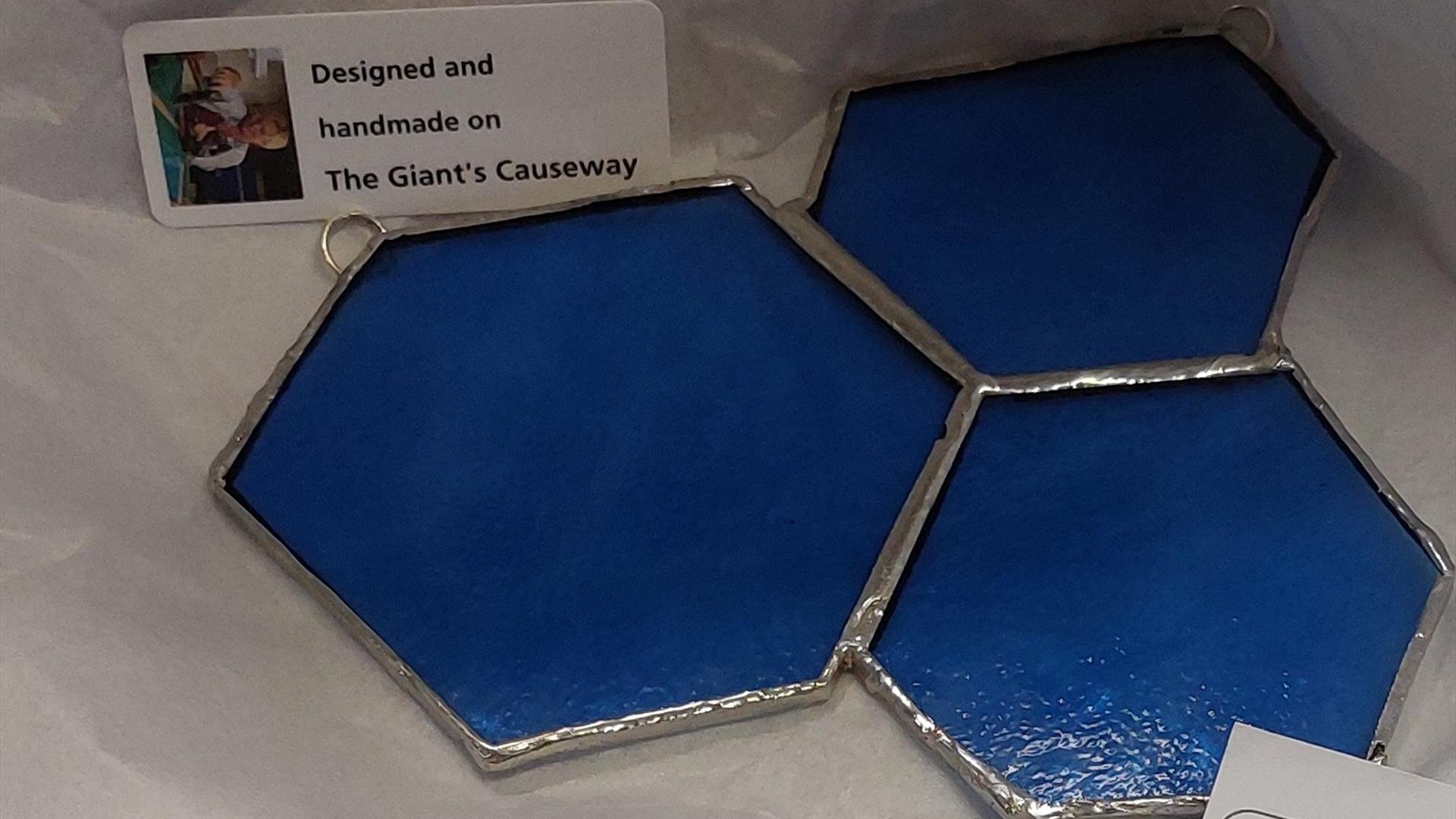 Image of 3 hexagonal shaped blue colour pieces of glass, inspired by the shape of the causeway stones at The Giant's Causeway. They are linked together by a silver colour solder to create a decorative hanging piece. Beside them is a white card with an image of the glass artist Eleanor-Jane McCartney and the words Designed and Made on The Giant's Causeway.